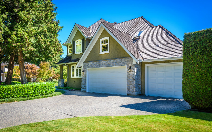 Large two-story suburban house with a gray shingle roof, green stucco and stone exterior, a wide concrete driveway, and a three-car garage under a clear blue sky.